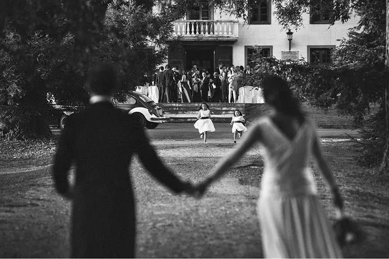 Novios de la mano mientras dos niñas corren hacia ellos frente a un palacete, reportaje de boda en blanco y negro de Josu Izarra
